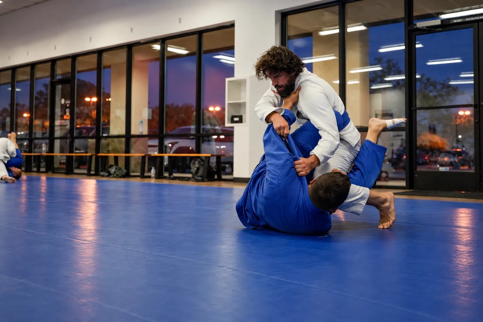 Two adults training on the mat in front of tall windows at dusk
