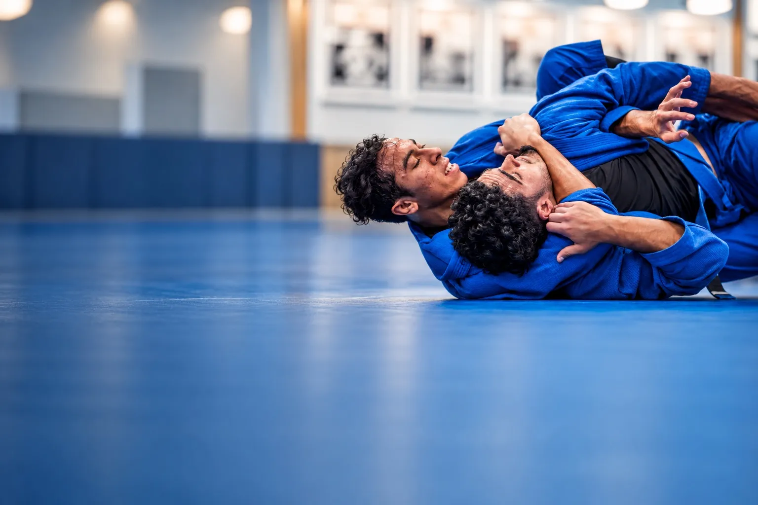 Two adults in blue gis training on the mat, both smiling mid-roll