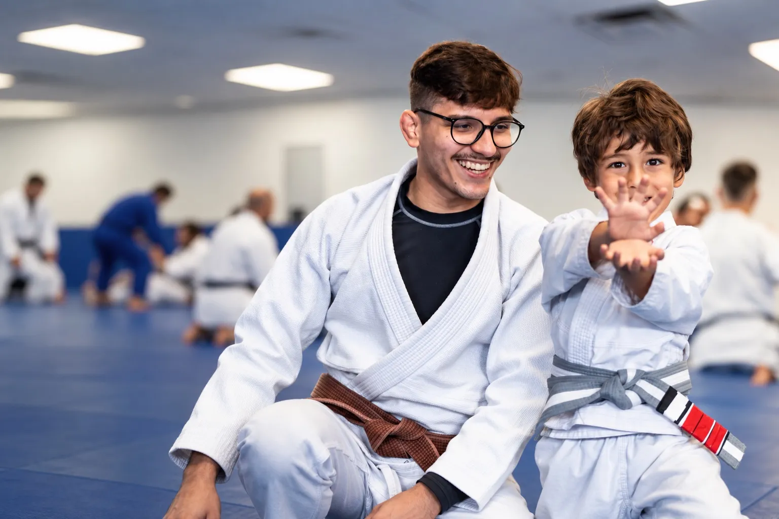 A smiling coach in a brown belt sits beside a laughing young student who reaches toward the camera